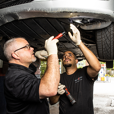Two auto repair technicians inspect the underside of a vehicle in Alexandria